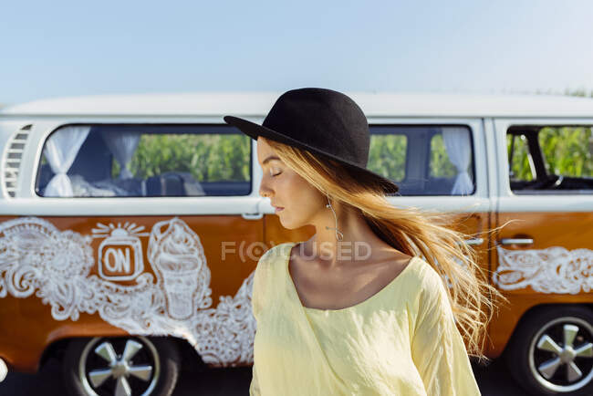 Linda chica rubia vestida con ropa de verano con un sombrero de pie fuera de una furgoneta - foto de stock