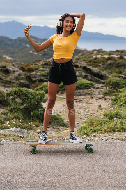 Pieno corpo di gioiosa femmina etnica in cuffie wireless in piedi su longboard e scattare autoritratto su smartphone in Cap de Creus — Foto stock