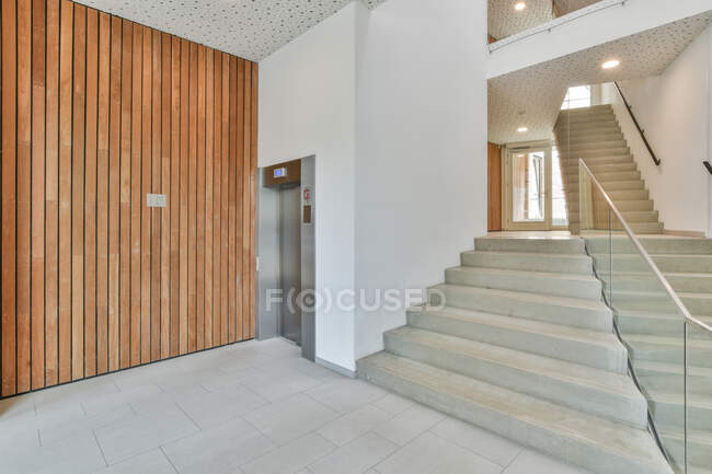 Интерьер просторного коридора нового жилого дома с деревянной стеновой лестницей и лифтом и встроенными лампочками на потолке — стоковое фото