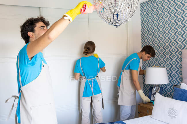 Positive young professional doctor cleaning staff in uniforms and gloves washing floor and wiping wardrobe and lamps during work in elegant bedroom — Stock Photo