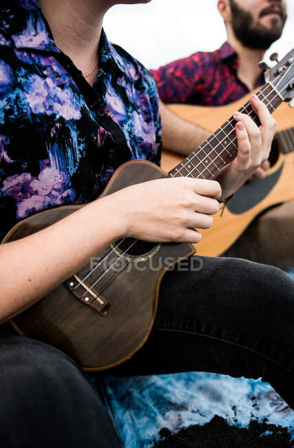 Cropped unrecognizable talented female musician with brown hair in casual clothes playing ukulele and singing song while sitting on sandy beach in nature in daylight — Stock Photo