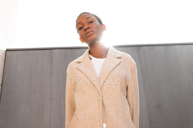Serious backlit African American female with short hair looking at camera while standing near wall on street with bright sunlight — Stock Photo