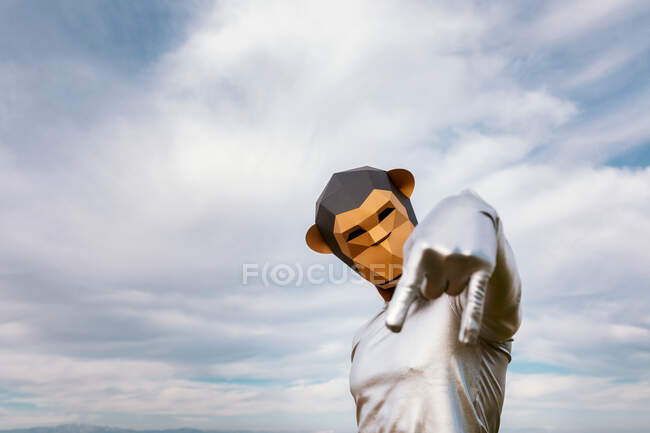 Pessoa auto-assegurada com máscara de macaco geométrico mostrando dedo indicador e rosa para baixo contra as nuvens — Fotografia de Stock