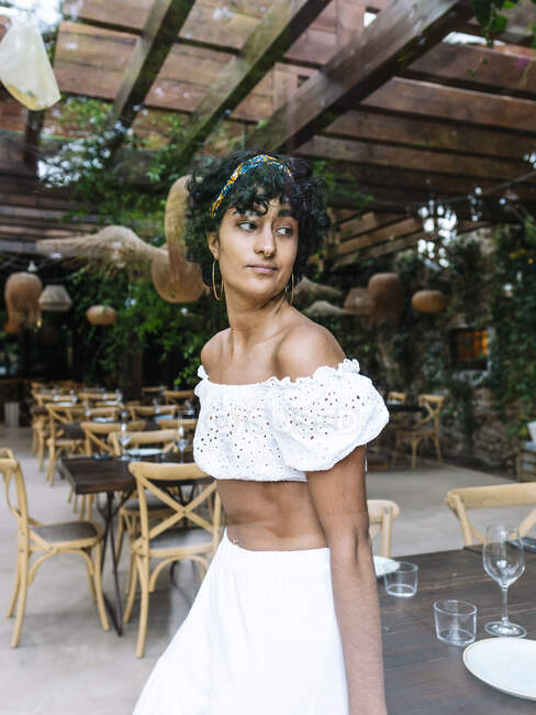 Durch das Fenster einer trendigen ethnischen Frau in weißem Outfit und mit lockigem Haar, die neben einem Tisch im Café steht und wegschaut — Stockfoto