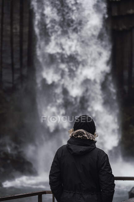 Vista posterior de un turista anónimo en ropa interior cálida y sombrero de pie en el mirador y admirando la impresionante cascada de Svartifoss durante el viaje a Islandia - foto de stock