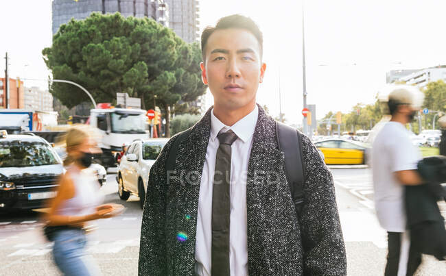 Auto assicurato giovane imprenditore maschio asiatico in elegante usura guardando la fotocamera contro le persone irriconoscibili nella città soleggiata — Foto stock