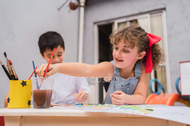 Crianças positivas com pincéis pintura com aquarelas coloridas sobre papel à mesa com suprimentos em sala de luz com quadro branco — Fotografia de Stock