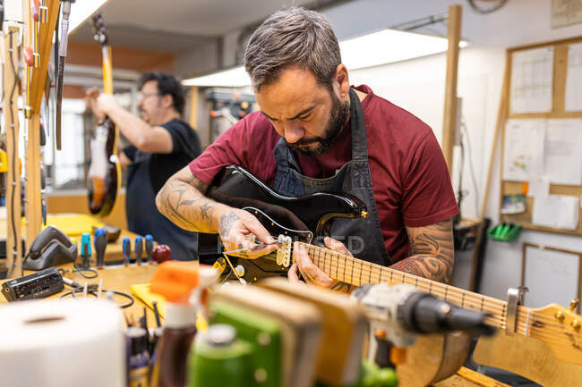 Professional master in apron standing near table with instruments and tools while tuning electric guitar in modern garage — Stock Photo