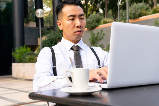 Wistful young Asian male entrepreneur with cup of hot drink and netbook looking at screen in urban cafeteria table in daylight — Stock Photo
