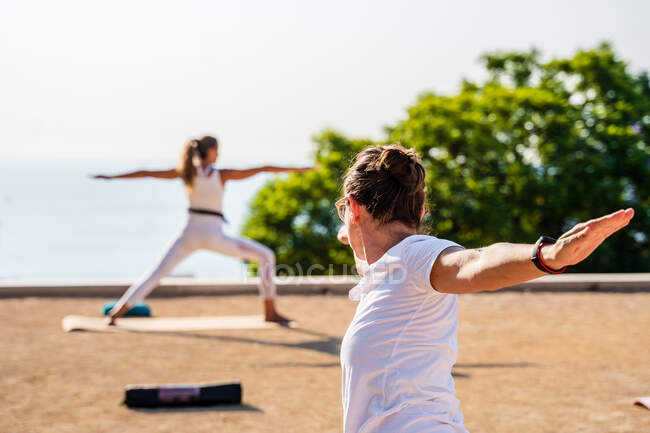 Unrecognizable women in white clothes standing with raised arms and performing Virabhadrasana B during yoga lesson in park in sunny day — Stock Photo