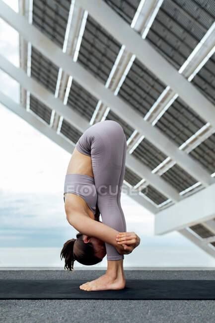 Повний вид збоку тіла спортивної жінки, що практикує балансування, стоячи вперед згинання асани, стоячи біля фотоелектричної панелі під час тренування йоги — стокове фото