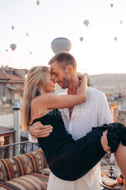 Man holding girlfriend in hands while standing with eyes closed on terrace on background of hot air balloons in sunlight — Stock Photo