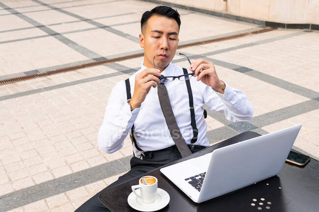 Хорошо одетый молодой этнический предприниматель мужчина надевает очки на стол с нетбуком и горячим напитком в уличной столовой — стоковое фото