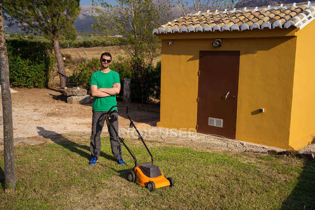 Young man in sunglasses standing with crossed arms near lawn mower and yellow building on grassy lawn in summer — Stock Photo