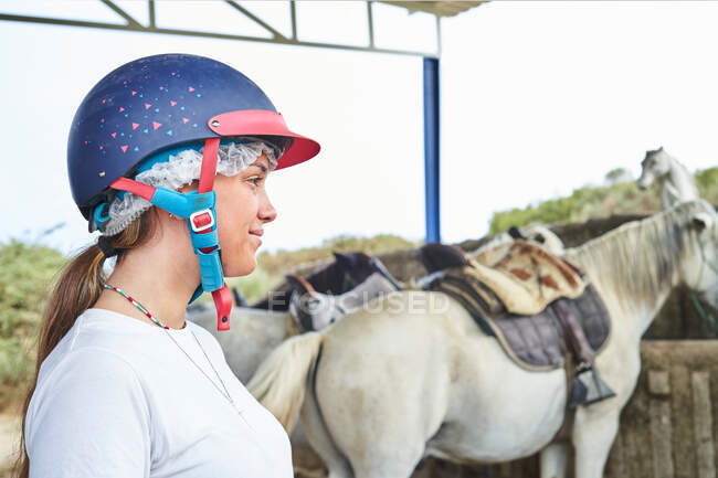 Chica adolescente en ropa casual con casco mientras está de pie cerca de caballos con sillas de montar en el patio de cultivo en establo durante el día - foto de stock