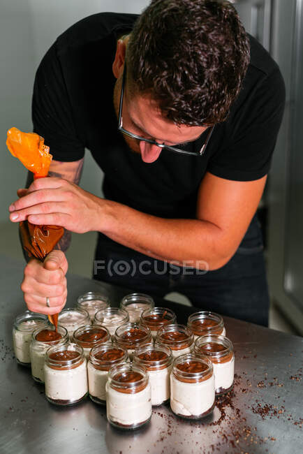 D'en haut de jeune boulanger masculin attentif en vêtements décontractés et lunettes ajoutant garniture de caramel de sac à pâtisserie dans des pots avec délicieux dessert couche dans la cuisine — Photo de stock