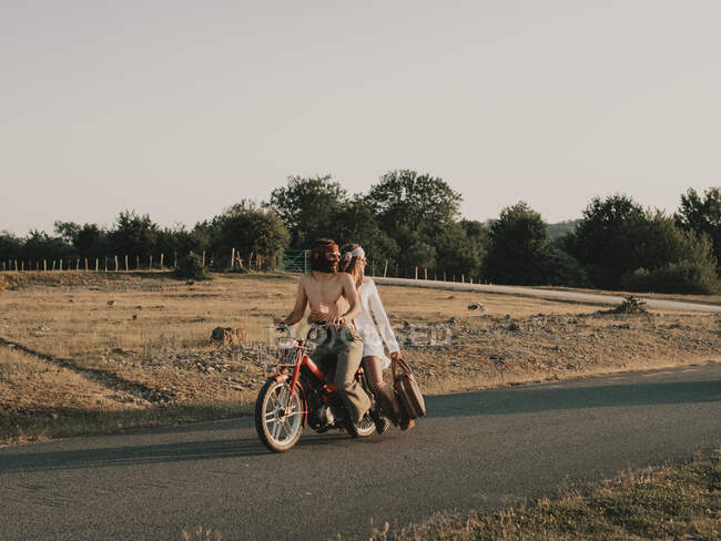 Full body of hippie couple riding on red moped on asphalt roadway during trip in nature with trees on summer day — Stock Photo