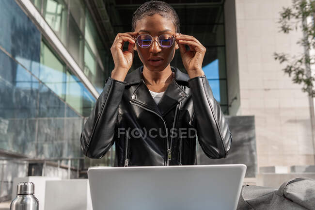 Desde abajo mujer afroamericana freelancer en chaqueta de cuero sentado con netbook en vueltas durante el trabajo en línea en la calle mientras se pone las gafas - foto de stock