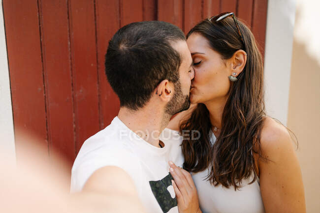 Вид сбоку на влюбленного молодого бородатого этноса, целующегося с девушкой, когда он делает селфи, стоя на улице в солнечный день — стоковое фото