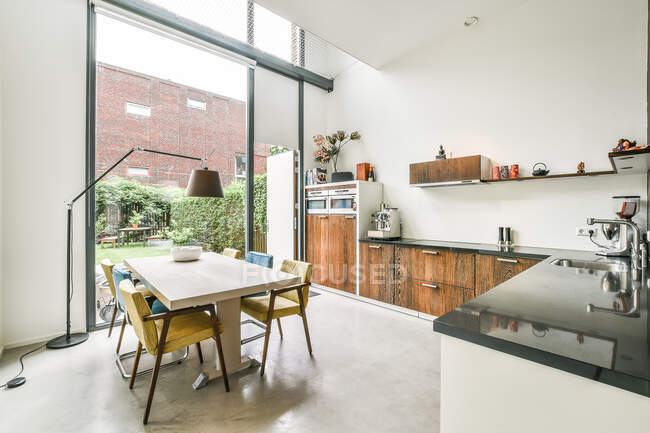 Sillas en la mesa colocadas cerca de armarios con varios electrodomésticos modernos en amplia cocina con puerta con vistas a patio con plantas verdes - foto de stock