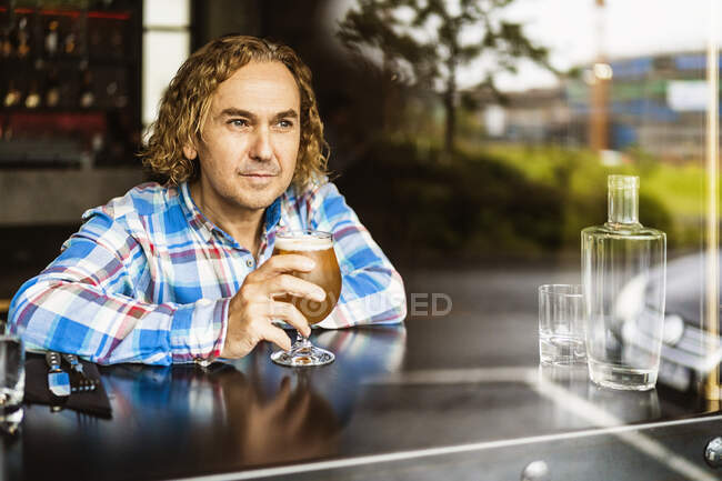 Nachdenklicher Mann mit lockigem Haar in lässiger Kleidung sitzt an Holztheke neben Fenster in Bar und trinkt tagsüber Bier — Stockfoto
