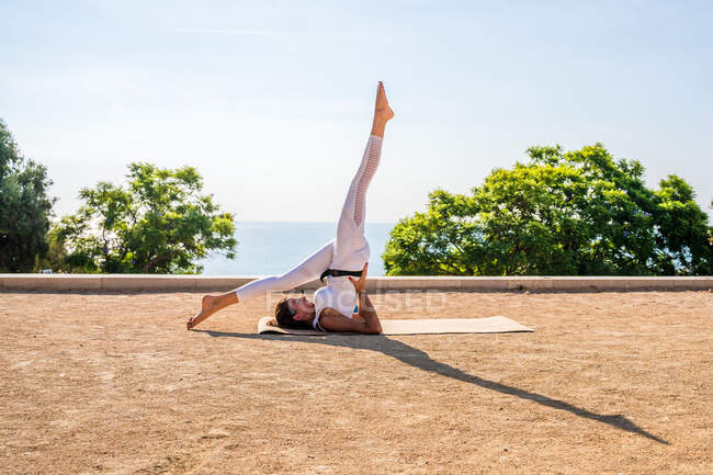 Vista lateral da fêmea flexível em activewear realizando Eka Pada Sarvangasana no tapete em solo seco durante sessão de ioga no parque contra árvores verdes e céu azul sem nuvens na luz solar — Fotografia de Stock