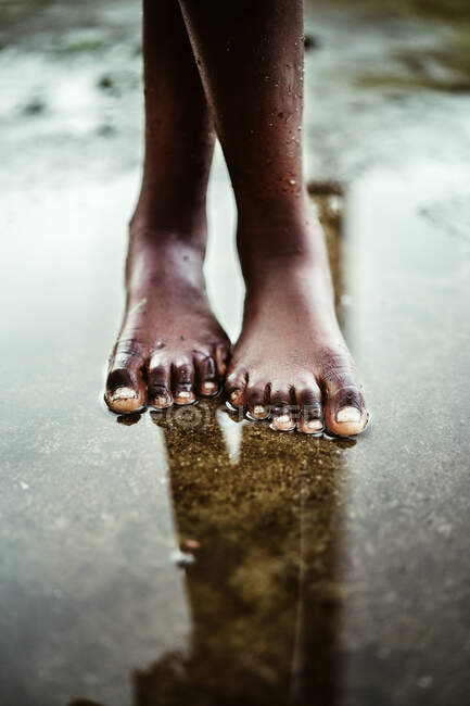 From above of crop unrecognizable black barefoot kid standing in small puddle on asphalt road on street on So Tom and Prncipe island in daylight — Stock Photo