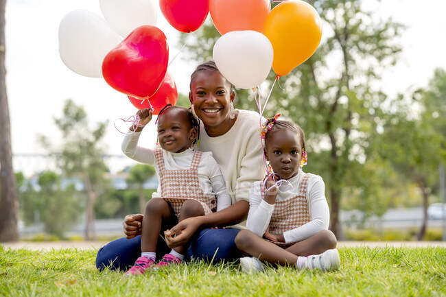 Счастливая афроамериканская мать сидит с позитивными маленькими дочерьми с красочными воздушными шарами на зеленой траве в парке и смотрит в камеру при дневном свете — стоковое фото