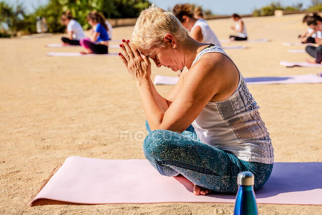 Спокійна жінка з коротким волоссям в активному одязі робить Падмасану руками Намасте, сидячи на йога маті під час тренувань на відкритому повітрі в сонячний день. — стокове фото