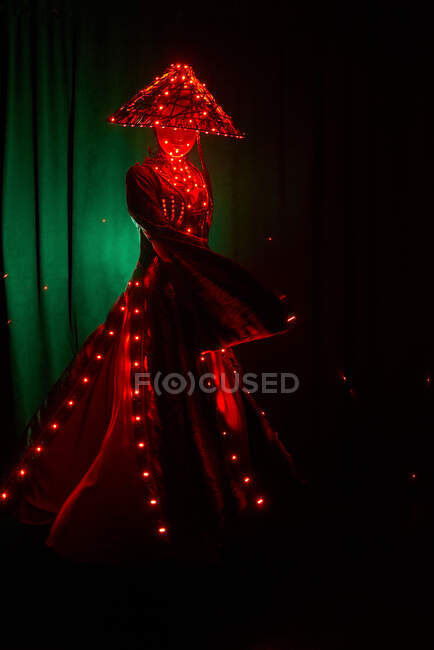 Mulher irreconhecível com uma máscara em roupa tradicional criativa e headwear vietnamita com iluminação vermelha em pé no estúdio escuro em fundo preto durante a performance — Fotografia de Stock