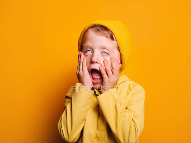 Little boy in trendy raincoat and beanie hat standing with opened mouth and covering eyes while touching cheeks and making grimace against yellow background in studio — Stock Photo