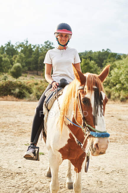 Full length of happy girl in helmet and casual outfit sitting in saddle on horse with bridle on sandy meadow near grass in nature in daylight — Stock Photo