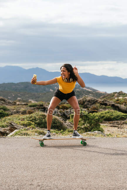 Full body of joyful ethnic female in wireless headphones standing sticking tongue out on longboard and taking self portrait on smartphone in Cap de Creus — Stock Photo
