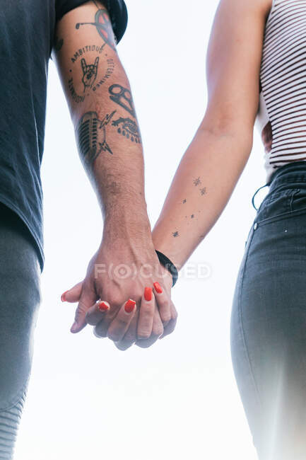 From below of crop unrecognizable couple with tattoos on arms in casual clothes standing and holding hands against light background in daytime — Stock Photo