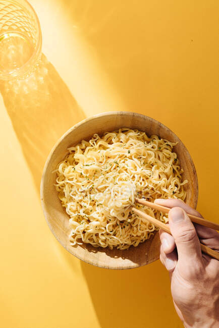 Top view of crop anonymous person with wooden chopsticks eating fresh noodles in bowl placed on yellow background with glass — Stock Photo