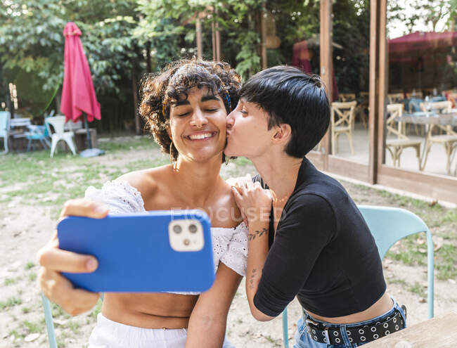 Content friendly multiracial females taking self shot on mobile phone enjoying weekend together in summer park while they kiss — Stock Photo