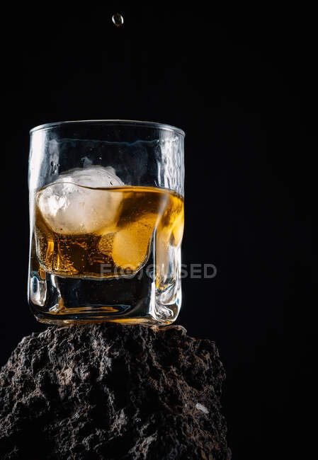 Gotas de uísque caindo em cubos de gelo servidos em vidro cristal colocado em superfície áspera contra fundo preto — Fotografia de Stock