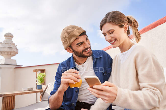 Mujer joven sonriente con vaso de agua sentado cerca de novio barbudo y el teléfono inteligente de navegación en el sofá en la terraza - foto de stock