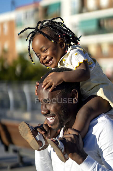 Happy African American girl with dark braids sitting on shoulders of cheerful father and jumping while having fun together on street in sunlight — Stock Photo