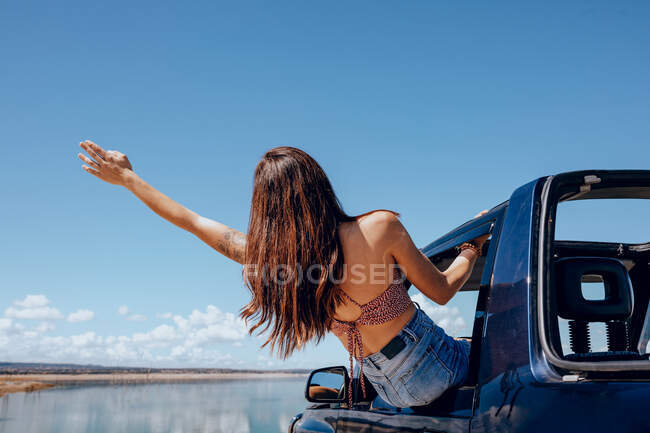 Back view of young anonymous female in denim jeans and top leaning out of car window and waving hand on coast of pond — Stock Photo