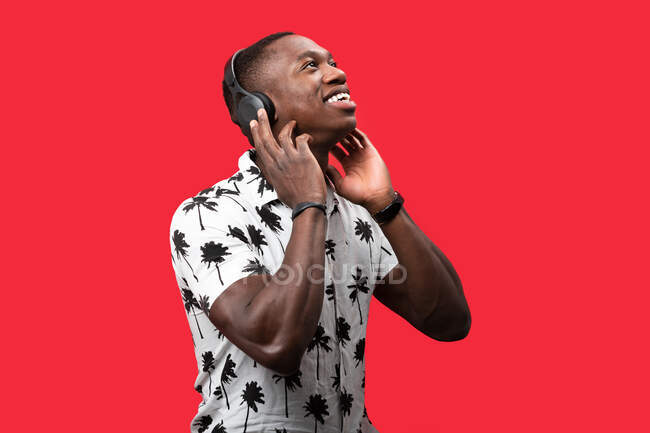 Glad African American male listening to songs in wireless headphones with pleasure while standing against red background and looking up — Stock Photo