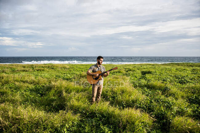 Calm man musician in casual clothes standing among green grass on coast of ocean and playing acoustic guitar in summertime in daylight — Stock Photo