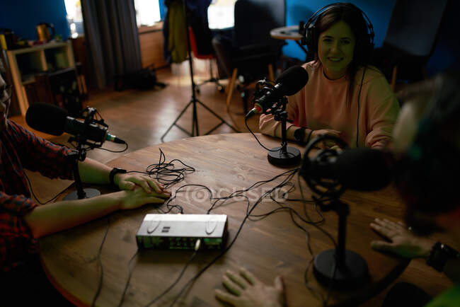 Young female radio host with headphones sitting at table with microphone and communicating with crop anonymous colleague during podcast recording in studio — Stock Photo