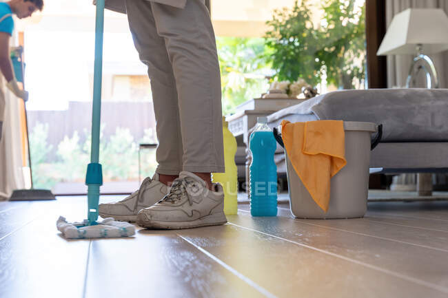 Zugeschnittene unkenntliche Kollegen in Schürzen und Handschuhen reinigen Parkettboden mit Mopp und Besen bei der Arbeit im modernen, geräumigen Wohnzimmer — Stockfoto