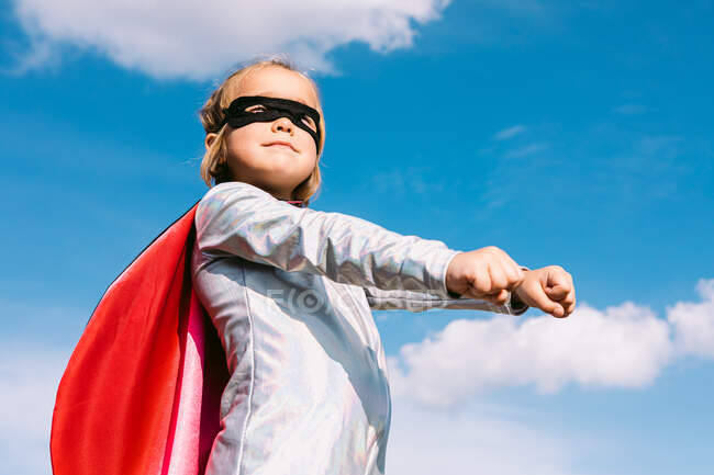 From below small girl in superhero costume raising outstretched fists for showing power while standing on rocky hill — Stock Photo