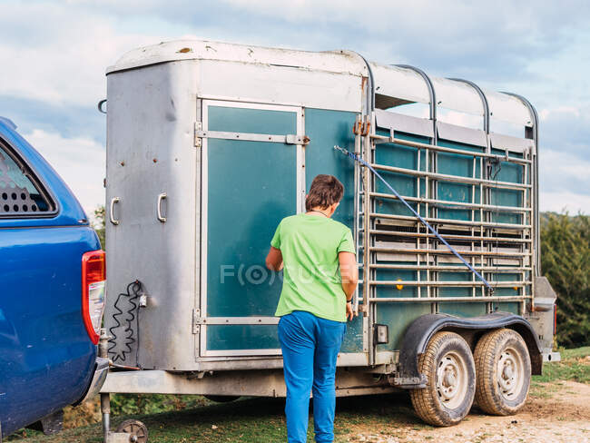 Back view of anonymous farmer in casual clothes closing door of livestock trailer attached to SUV car on rough road under cloudy sky in countryside — Stock Photo