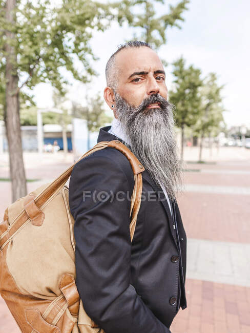 Vista lateral de homem barbudo sério em desgaste formal de pé olhando para a câmera perto da estrada na rua com edifícios modernos — Fotografia de Stock
