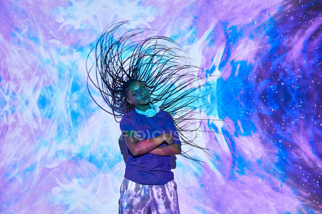 Vista lateral da fêmea étnica milenar sacudindo longos cabelos trançados enquanto seus braços são cruzados perto da parede com iluminação azul brilhante e lilás — Fotografia de Stock