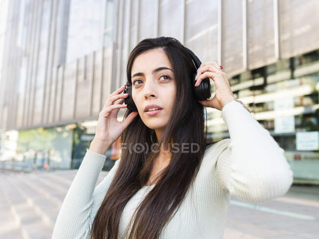 Calm woman with long brown hair in casual clothes adjusting headphones and looking away on street against modern building in city in daylight — Stock Photo