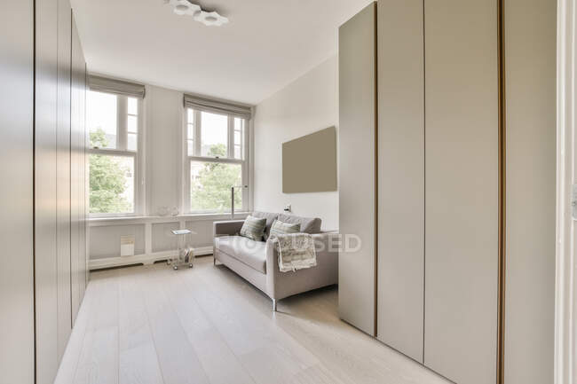 Sofá confortável com almofadas e roupeiros de estilo minimalista localizado perto de janelas na sala de estar iluminada pelo sol — Fotografia de Stock